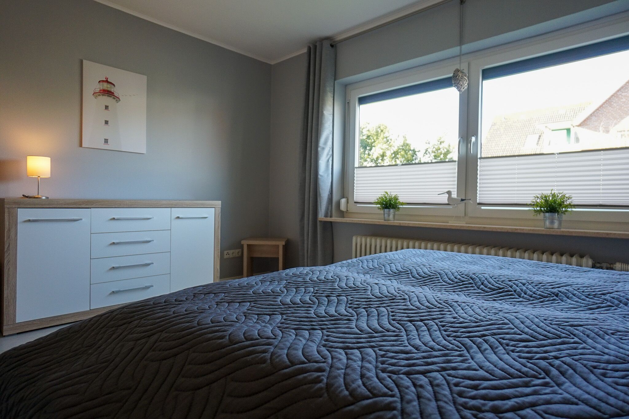 Um quarto com cama, penteadeira, janela com persianas e uma pequena mesa com abajur.