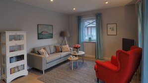 Ein Wohnzimmer mit grauem Sofa, einem roten Sessel, einem weißen Bücherregal, einem kleinen runden Tisch mit Lampe und einem Fenster mit Vorhängen.