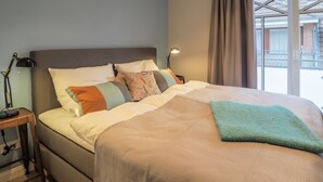 Ein ordentlich bezogenes Bett mit einer beigen Tagesdecke und mehreren Kissen, einer Nachttischlampe und Blick ins Freie durch ein Fenster.