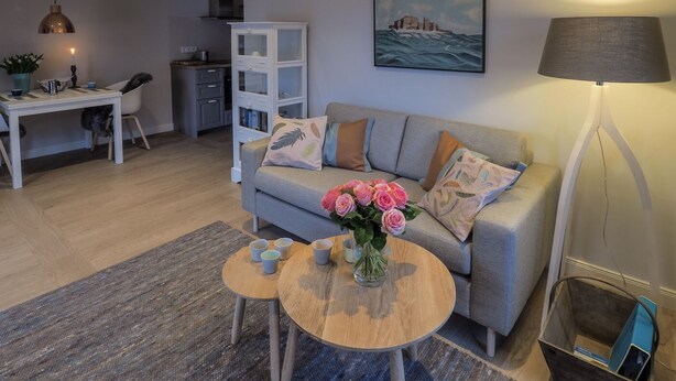 Ein modernes Wohnzimmer mit grauem Sofa, einem Couchtisch und einem Essbereich mit weißem Schrank und einer Pendelleuchte.