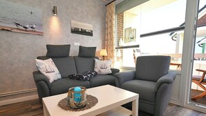 Een moderne woonkamer met een grijze bank, een bijpassende fauteuil en een witte salontafel. Op de bank staat een glazen tafellamp, en aan de muur hangt een ingelijst kunstwerk.