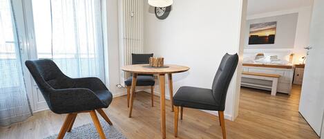 A modern living room with a round wooden table, two black chairs, a grey rug, and a wooden floor.