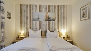 A double bed with white linens, two bedside tables with lamps, a framed picture of a sailboat on the wall, and a small framed artwork on the right. - Seemeile 2.18 Seestern (Büsum)