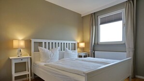 Et soveværelse med en hvid seng, to sengeborde, et vindue med gardiner og en radiator.