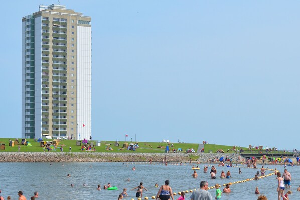 A high-rise building with multiple balconies overlooking a beach area with people swimming and sunbathing.