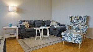En stue med en grå sofa, et hvidt sofabord, et lille rundt bord, en mønstret lænestol og en gulvlampe.