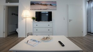 A modern living room with a flat-screen TV, a dining table with a bowl of fruit, a remote control, and a lamp. - Wohnung Deich Mitten in Büsum (Büsum)