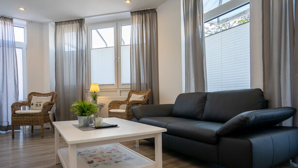 A living room with a black leather sofa, two wicker chairs, a white coffee table with a plant and a remote control, and a window with curtains. - Wohnung Deich Mitten in Büsum (Büsum)