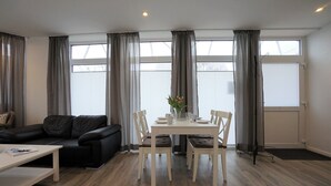 A modern living room with a black leather sofa, a white dining table with four chairs, and a coffee table. There are large windows with grey curtains, and a black floor lamp is visible. - Wohnung Deich Mitten in Büsum (Büsum)