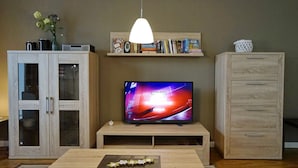 A living room with a flat-screen TV on a wooden stand, a coffee table with a decorative item, a bookshelf with books and decorative items, and a cabinet with drawers.