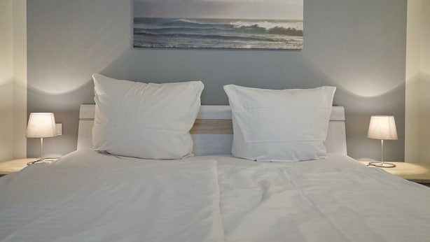 A neatly made bed with white linens, two pillows, and a wall-mounted picture of a beach scene.