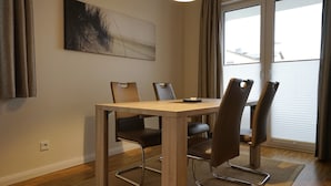 A dining area with a wooden table and four chairs, a painting on the wall, and a window with curtains.