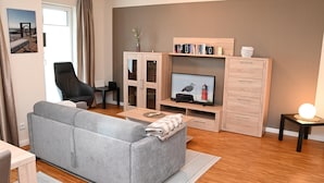 A living room with a grey sofa, a television, a wooden cabinet, and a small table with a lamp.