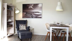 Una sala de estar moderna con un sofá gris, una mesa de comedor con sillas blancas, una lámpara colgante blanca y un cuadro con un paisaje en la pared.