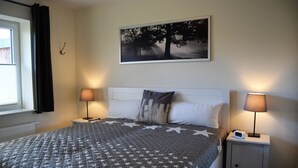 Una cama bien hecha con una colcha de diseño de estrellas, dos lámparas de noche, una mesita de noche con un reloj y un cuadro con un árbol en la pared.