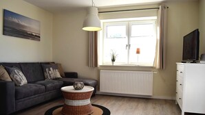 Una sala de estar con sofá gris, una mesita de mimbre, un colgante de techo, un radiador blanco y una ventana con cortinas.