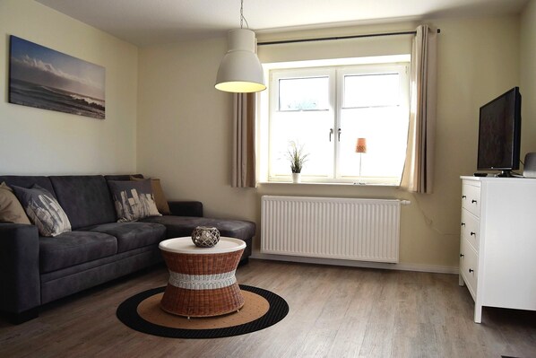 A living room with a grey sofa, a wicker side table, a hanging light fixture, a white radiator, and a window with curtains.
