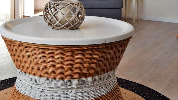 Una mesa de mimbre con superficie blanca, una silla de mimbre con un cojín estampado en forma de estrella, una lámpara fijada a la pared y un cuadro con una escena de playa.
