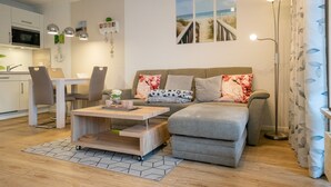 Een moderne woonkamer met een bank, salontafel en een eethoek. Achtergrond: een keuken met een magnetron en kastjes.