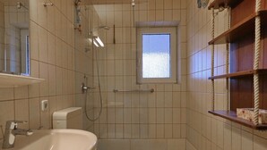 Un baño pequeño con inodoro, lavamanos y ducha. Hay una ventana sobre la ducha y un estante de madera con artículos de aseo.