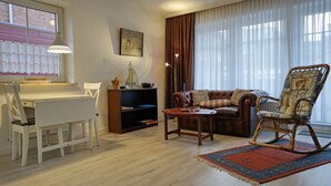 Una sala de estar con piso de madera, un escritorio blanco con una silla, un sofá marrón con cojines, una mecedora, una mesita y una estantería.
