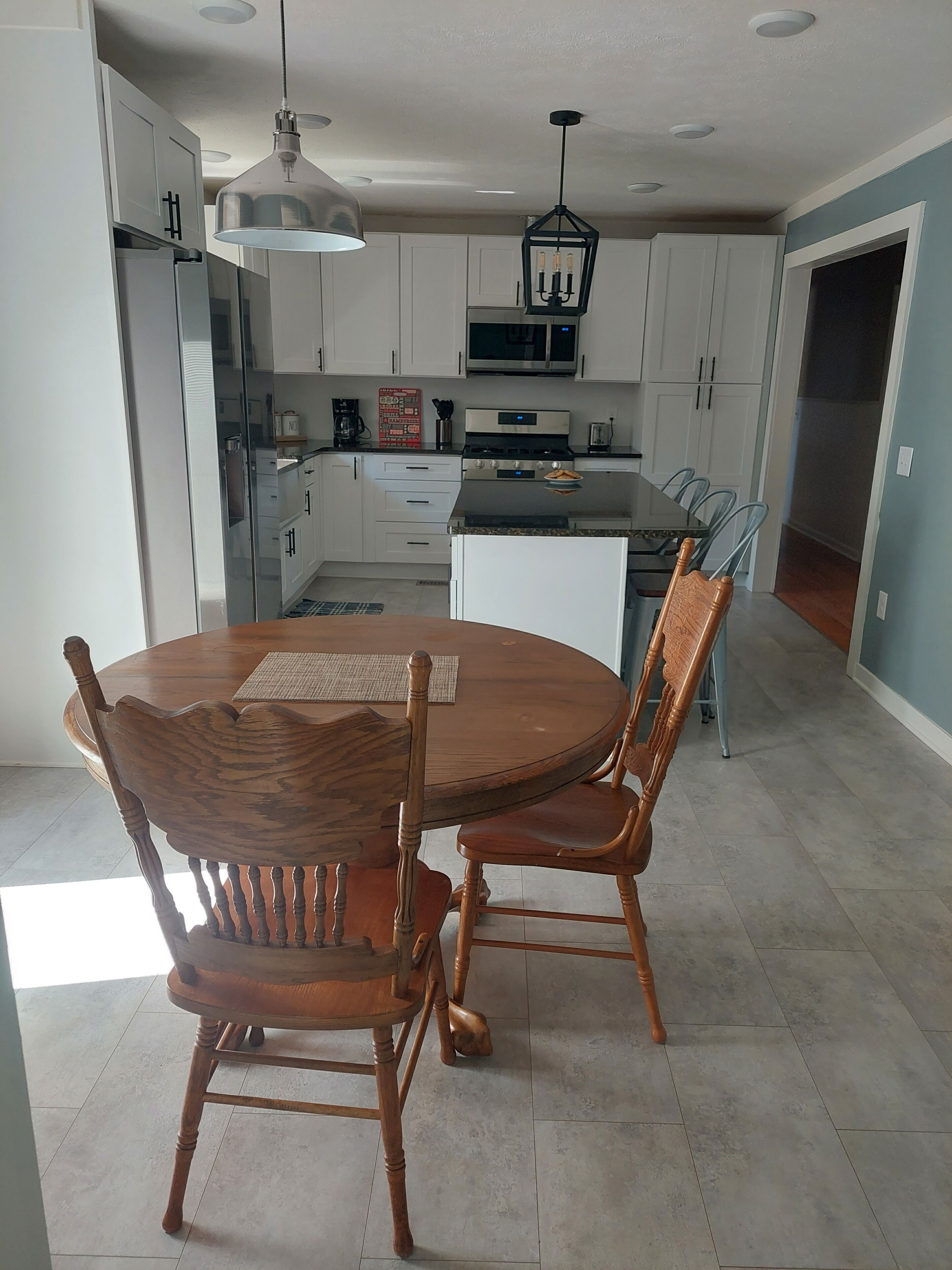 A modern kitchen with white cabinets, a stainless steel refrigerator, a microwave, and a gas stove. There is a dining table with four wooden chairs and a hanging pendant light.