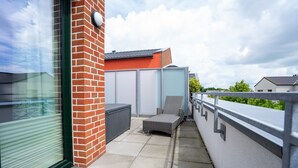 A balcony with a glass door, a brick wall, a grey outdoor chair, and a white wall with a window. - Wohnung Helios im Herzen von Büsum (Büsum)