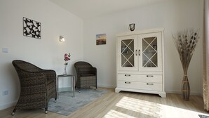 Ett rum med två fåtöljer i rotting, ett vitt skåp med glasdörrar, ett litet bord med en vas och en hög vas med torkade växter.