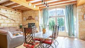 Un salon chaleureux avec un mur en pierre, des poutres apparentes, un canapé, une table à manger avec des chaises et une vue dégagée sur l’extérieur grâce à une grande fenêtre ornée de rideaux à carreaux.