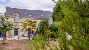 Un bâtiment à deux étages, doté d’un toit en pente, d’une façade en pierre et d’un petit jardin agrémenté de palmiers et de plantes en pot.