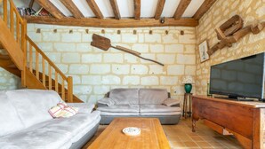 Un salon chaleureux avec un mur en pierre, des poutres apparentes, un canapé gris, une table basse en bois et un téléviseur posé sur un meuble en bois.