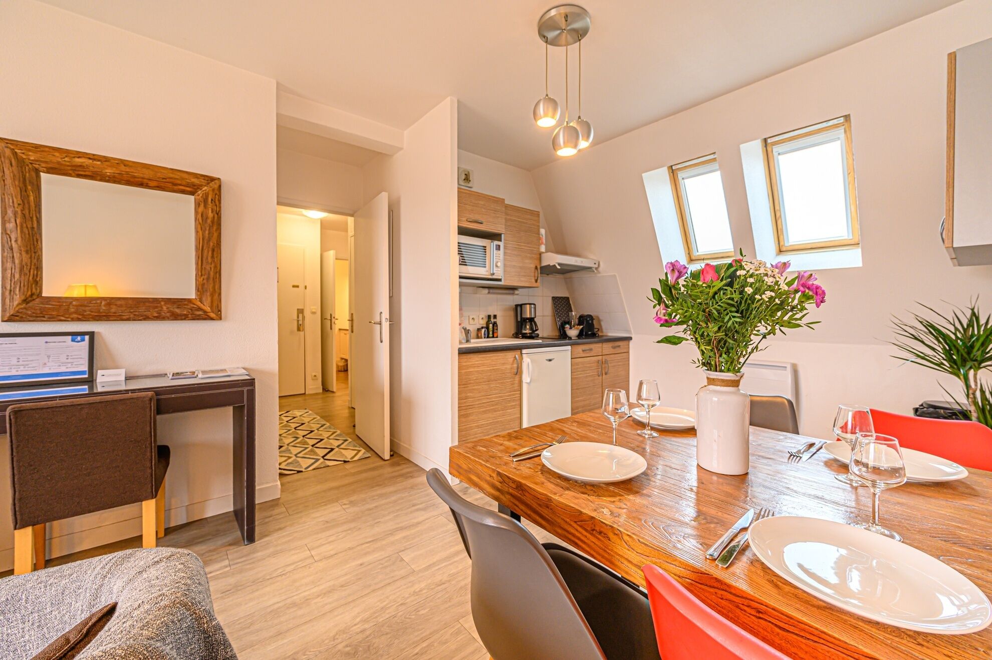 A modern kitchen with wooden cabinets, a dining area with a wooden table set for two, and a large mirror on the wall.