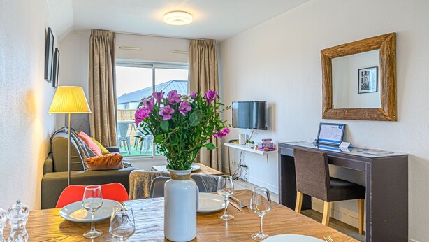 A hotel room with a dining table set for two, a TV, a desk with a computer, a large window with curtains, and a vase with flowers. - Le City-Break - T3 en centre-ville avec un parking sécurisé (Vannes)