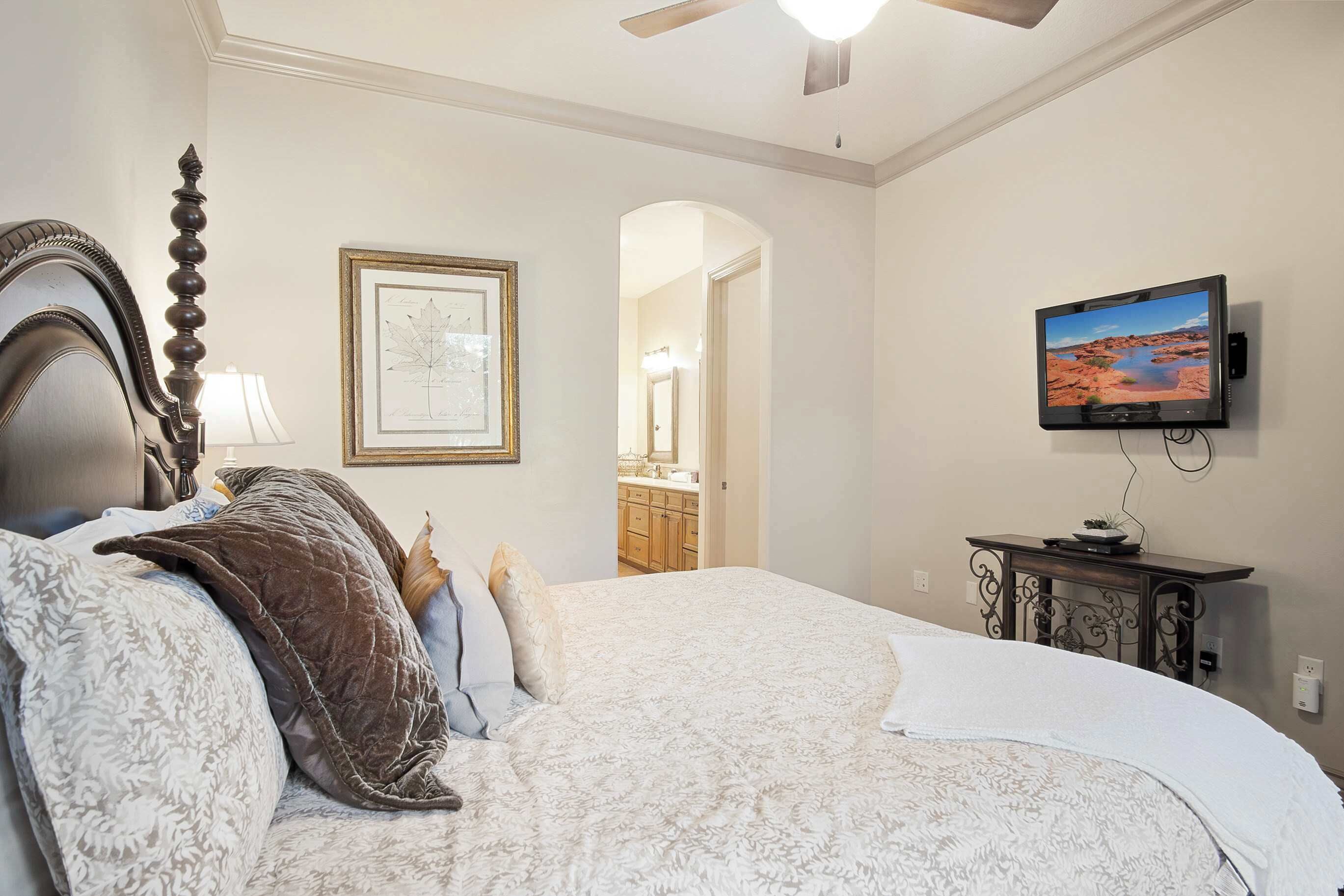 A bedroom with a large bed, a television mounted on the wall, a ceiling fan, and a framed botanical print on the wall.