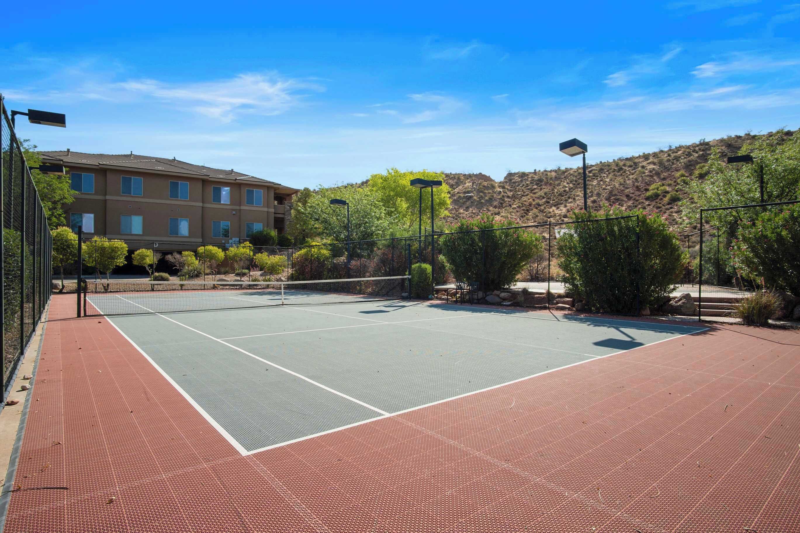 An outdoor tennis court with a net, surrounded by a fence and trees.