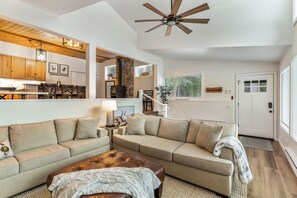 A spacious living area with a large sofa set, a coffee table, and a ceiling fan. A kitchen with wooden cabinets and a dining area with a table and chairs are visible in the background.
