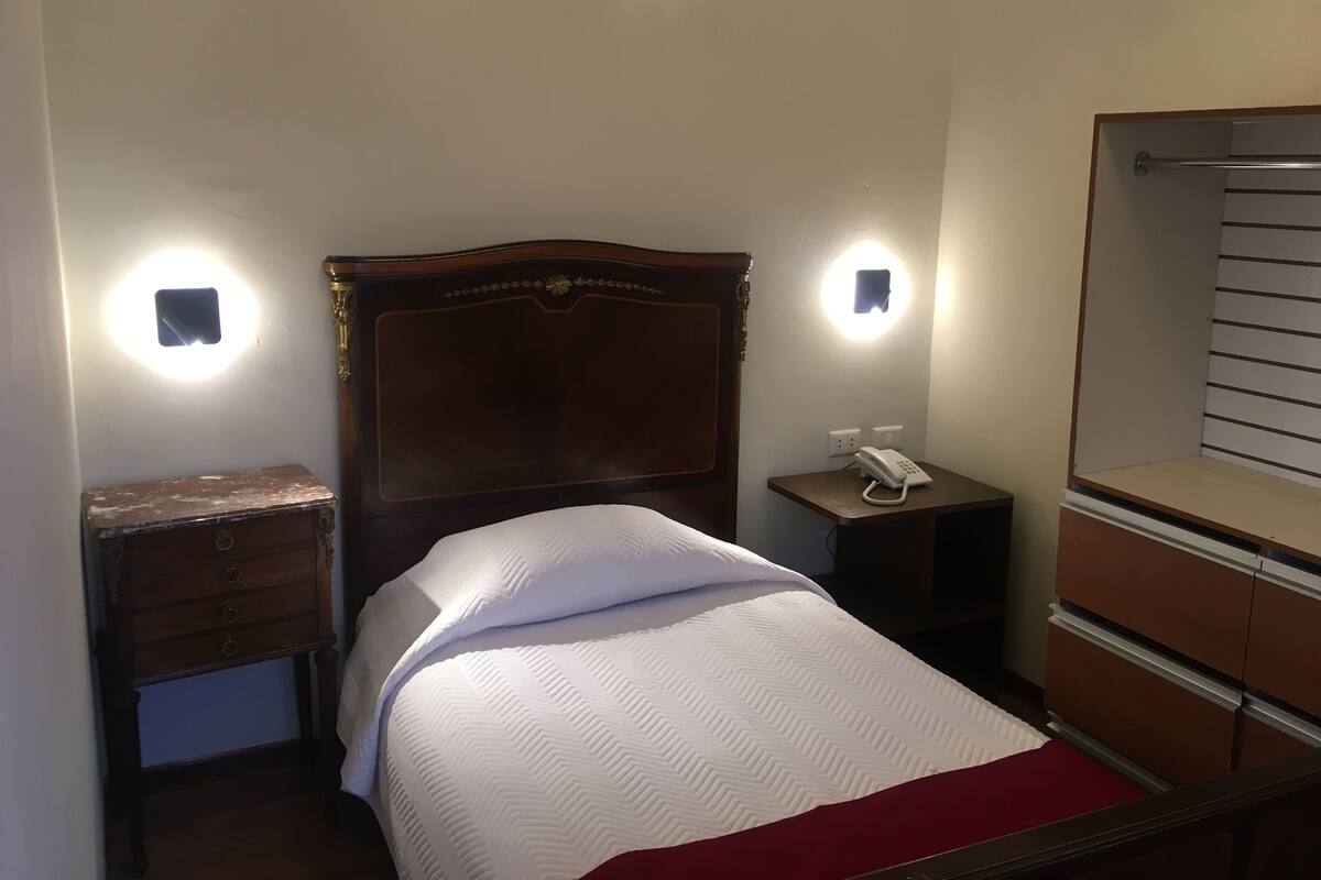 Una habitación de hotel con cabecera de madera, una mesita de noche con teléfono, una mesita auxiliar, un aparador y dos luces fijadas a la pared.