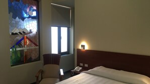Habitación de hotel con una cama, una silla, una mesita de noche, un teléfono, una ventana con luz natural y un vitral.
