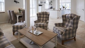 A spacious living area with a dining table, two armchairs, and a sofa. A kitchen is visible in the background with white cabinets and a large window. - Dreamlike vacation under thatch - Reethus "de Tweeling" Apartment ground floor (Büsum)