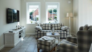 Un salon comprenant un canapé, un pouf et un fauteuil, une table basse, un téléviseur fixé au mur et une fenêtre donnant sur une maison.