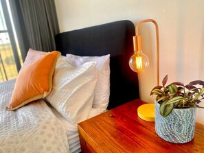 Ein ordentlich bezogenes Bett mit Kissen, ein Nachttisch mit Lampe und ein Blumentopf.