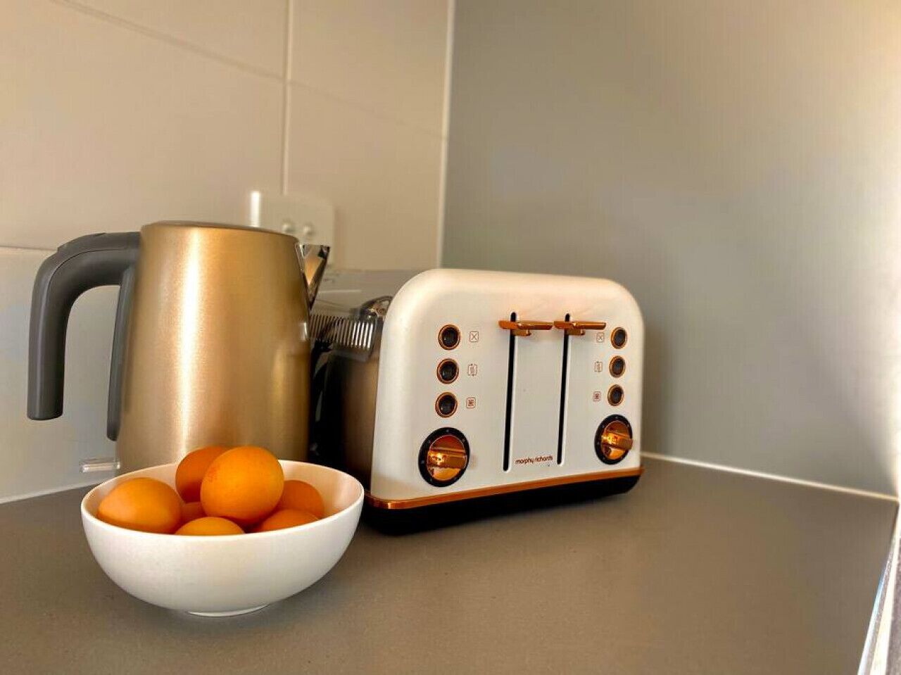 A kitchen counter with a white toaster, a golden kettle, and a bowl of oranges.