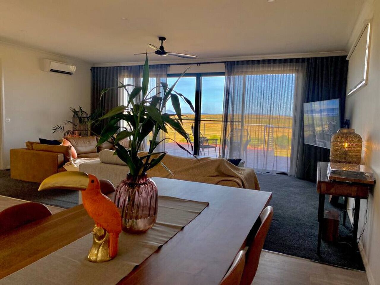 A modern hotel room with a dining area, a large window with a view, a ceiling fan, and a plant on the table.