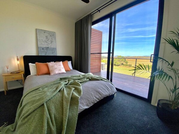 A bedroom with a large bed, a nightstand, a window with a view of the outdoors, and a potted plant.