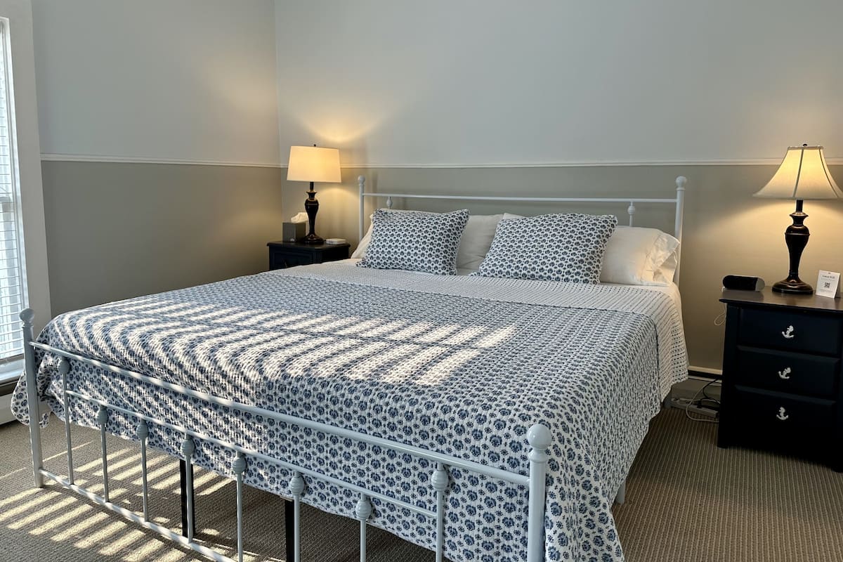 A neatly made bed with a patterned bedspread, two bedside tables with lamps, and a window with blinds.