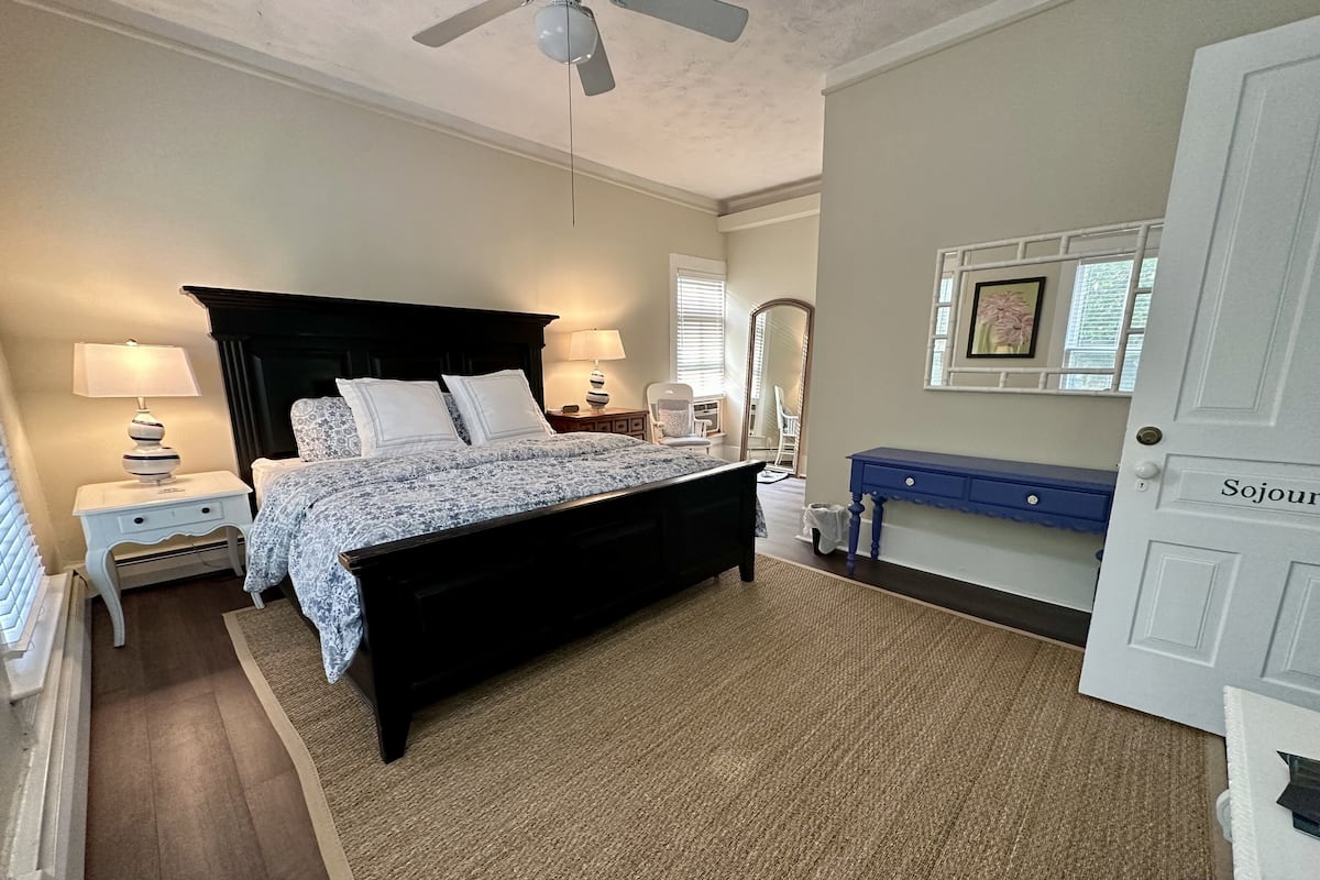 A bedroom with a bed, two bedside tables, a nightstand, a ceiling fan, a blue console table, and a door labeled "Sojour".