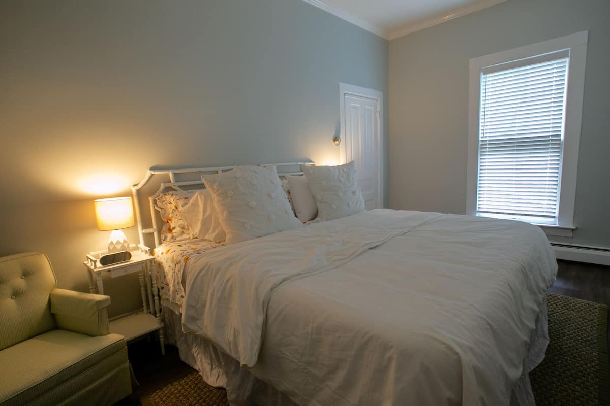 A bedroom with a bed, nightstand, lamp, chair, and window with blinds.