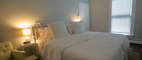 A bedroom with a bed, nightstand, lamp, chair, and window with blinds.