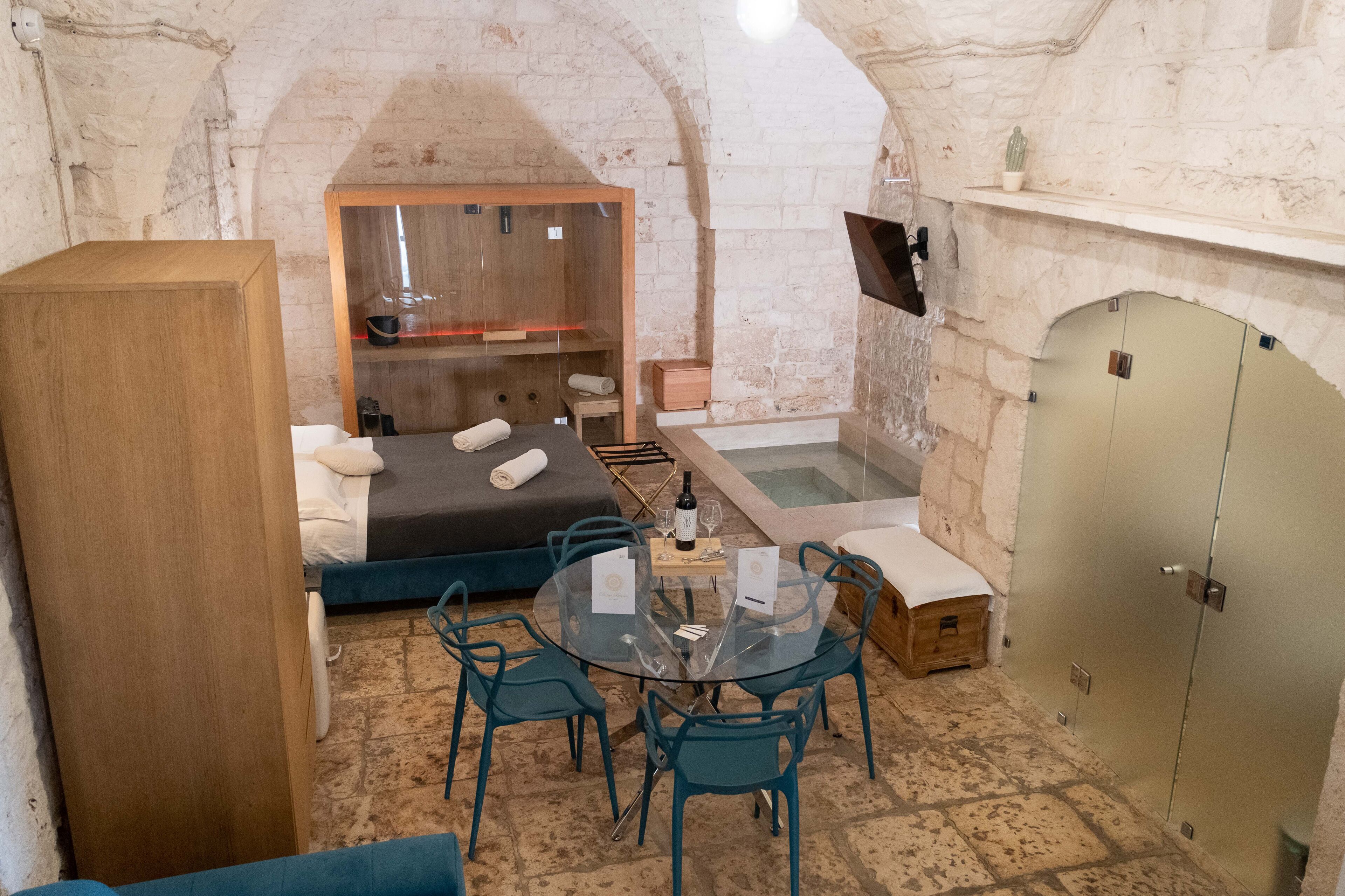 A cozy room with a bed, a small table with chairs, a glass-top table with a bottle and glasses, a wooden cabinet, and a stone wall.
