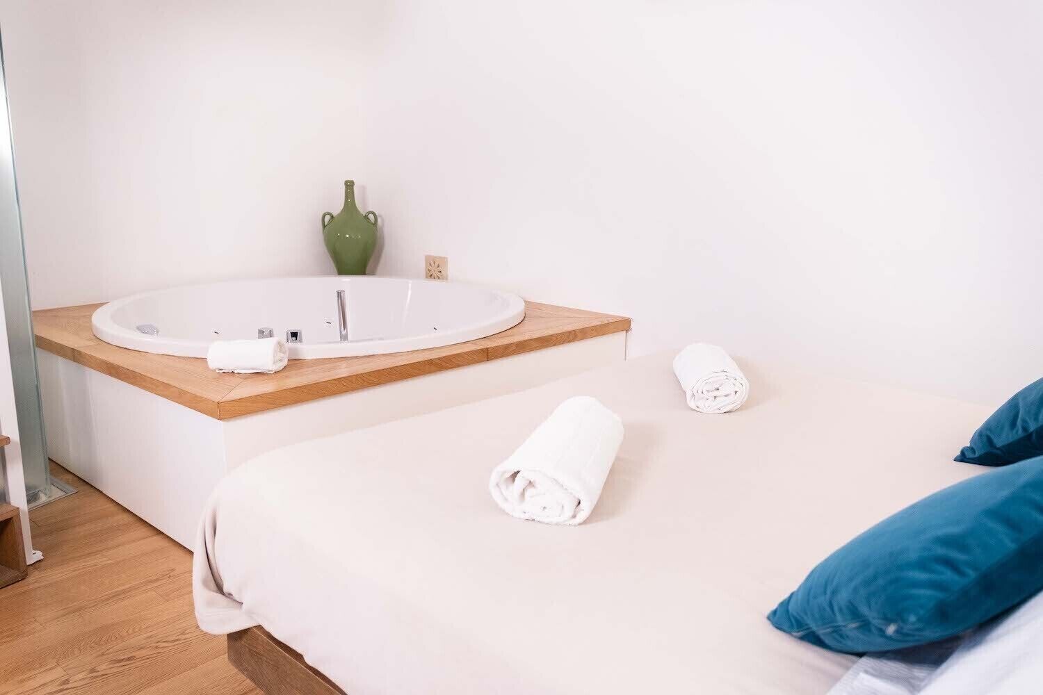 A bedroom with a bed, a wooden bedside table, a round white bathtub, and a green vase on the wall.